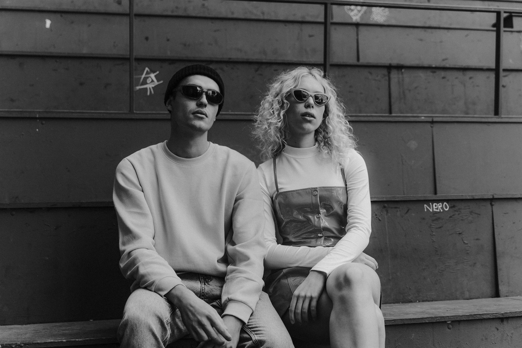 Two friends in sunglasses sitting side by side on a bench, black and white